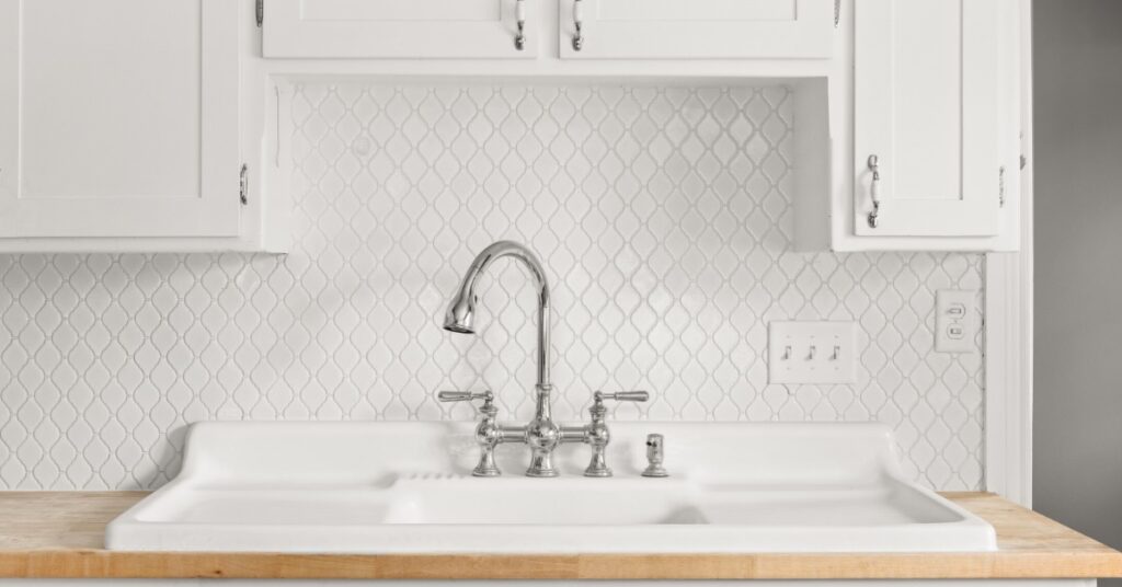 A kitchen faucet above a white farmhouse sink with double drainboards set against patterned tile and a wood countertop.
