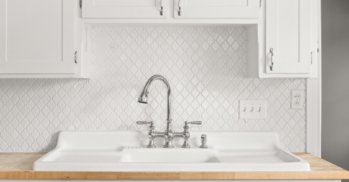 A kitchen faucet above a white farmhouse sink with double drainboards set against patterned tile and a wood countertop.